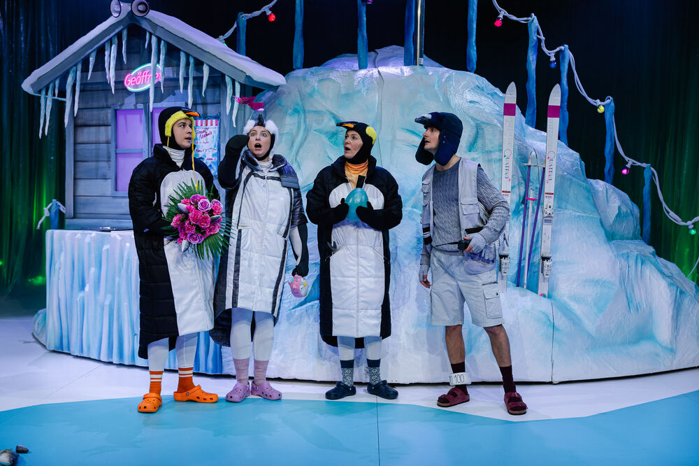 Vier Menschen stehen auf einer winterlich gestalteten Theaterbühne.
Drei von ihnen tragen Pinguin-Kostüme – lange weiß-schwarze wattierte Overalls mit Pinguinhauben – und bunte Crocs. Eine Person hält einen Strauß pinker Rosen, eine andere zeigt überrascht nach oben, die dritte hält ein türkisfarbenes „Ei“. Rechts steht eine weitere Person in einem Expeditions-Outfit mit grauem Strickpullover, kurzer Hose, Wintermütze und Sandalen mit Socken. Hinter ihnen befindet sich eine künstliche Eislandschaft mit einer kleinen Holzhütte, Eiszapfen, bunten Lichterketten und zwei Skiern.